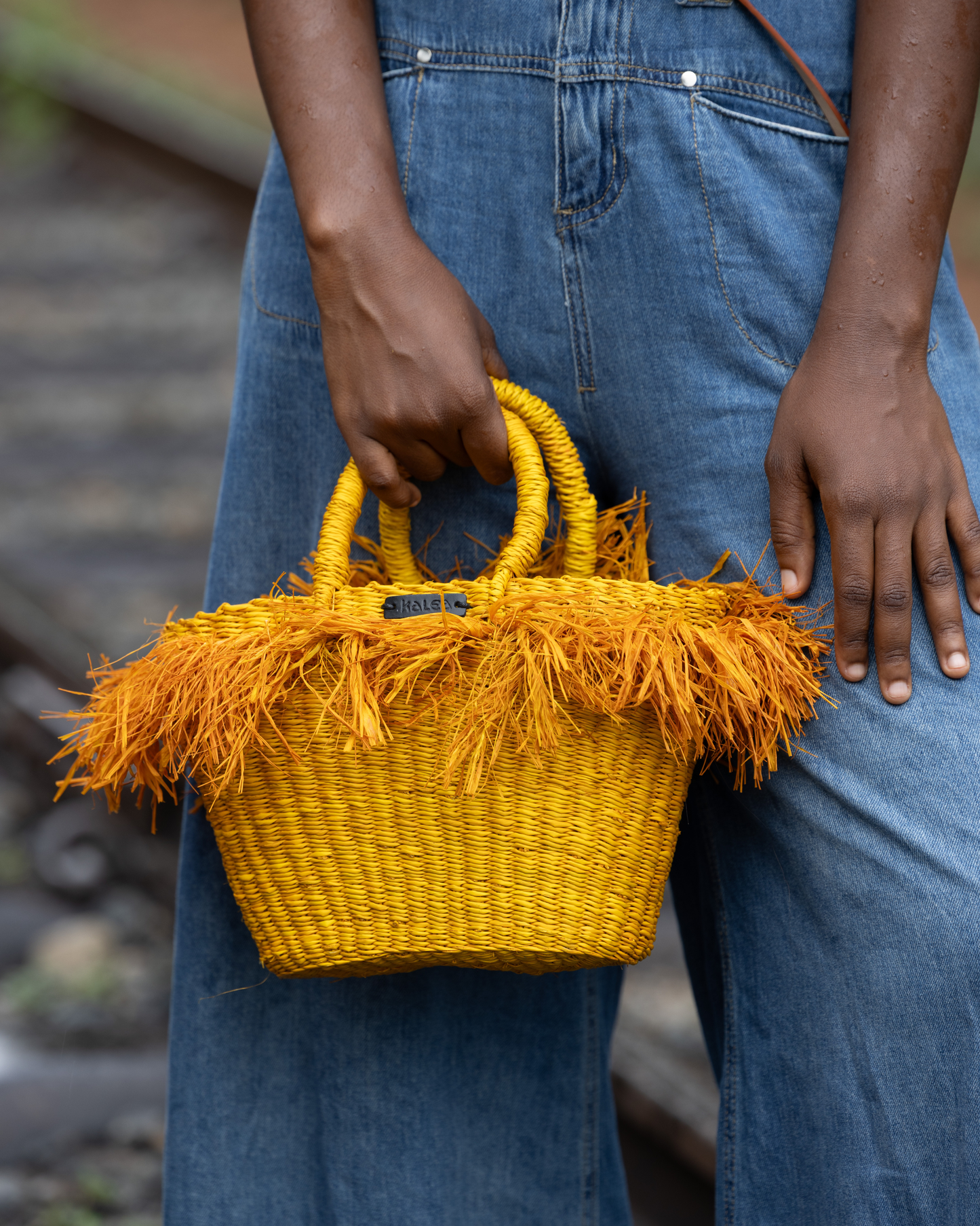 Person holding a yellow woven handbag with fringes, wearing blue jeans.