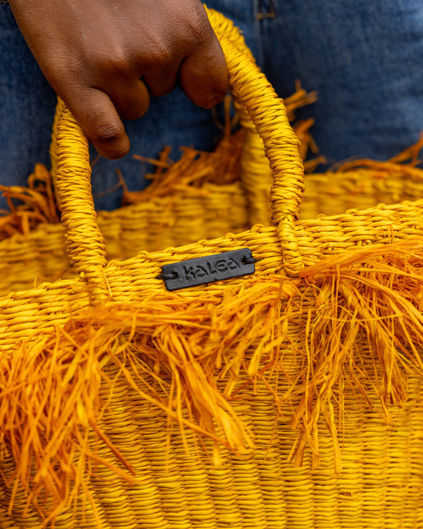 Yellow woven bag with 'Kales' branding held by a hand against a blue denim background